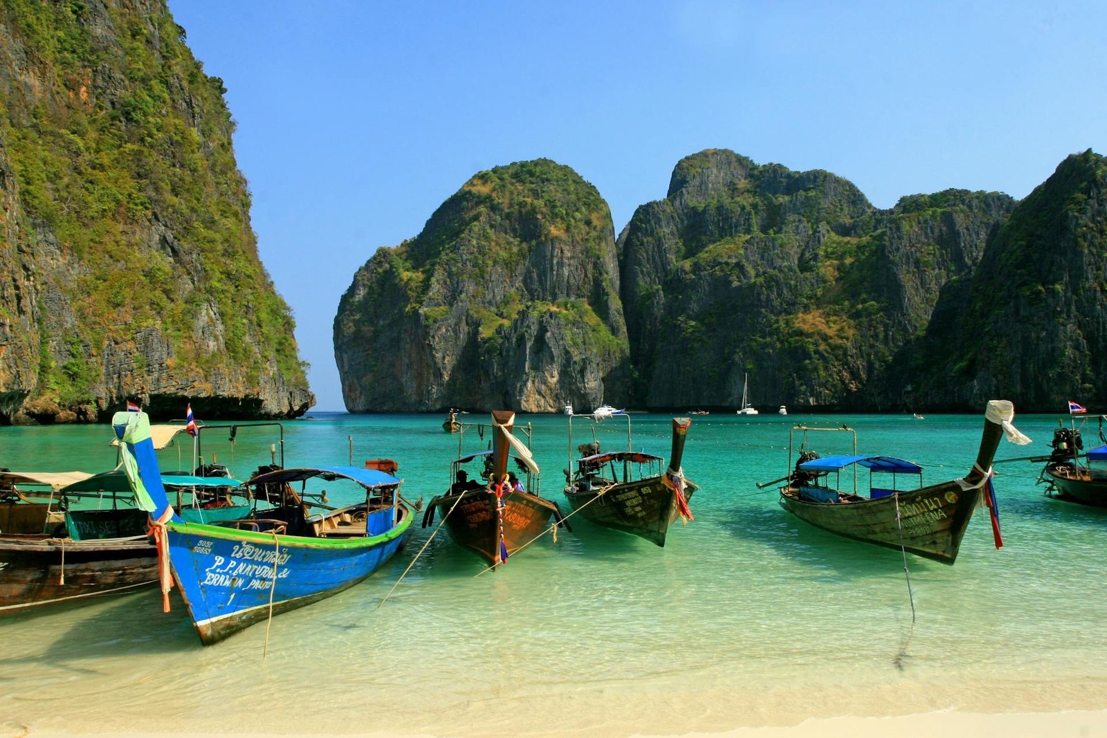 Traditional wooden boats at Maya Bay in Thailand, with limestone cliffs and crystal-clear water under a bright blue sky.